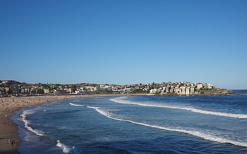 The full crescent of the beach seen from the southern end, the sand curving away toward the northern headland with low apartment buildings stepped up the hill above it.