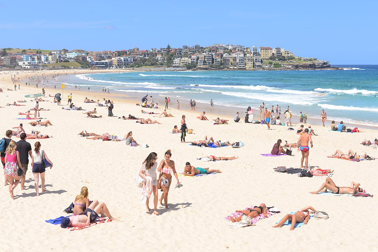 Sunbathers and swimmers spread across the sand at the height of the day, apartment blocks on the northern headland visible above the surf.
