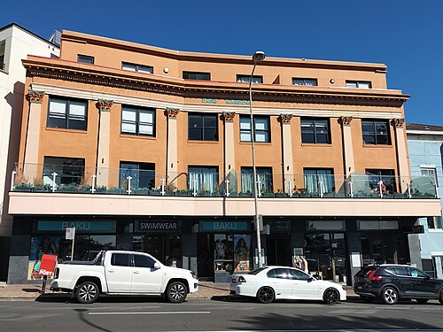 A three storey rendered building on Campbell Parade, orange paint over pilasters and ground floor shopfronts along the kerb.
