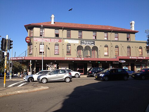 A corner pub building set back from the beach, beige plaster walls, a red tile roof and a wide awning over the footpath.