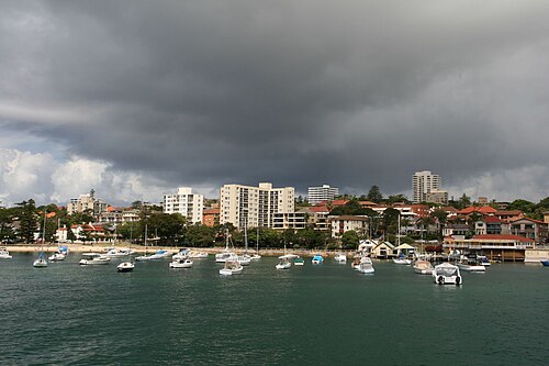 A wide view across the harbour, yachts moored off the shore and waterfront apartment buildings under a dark sky.