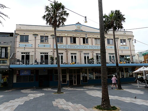 A three storey rendered corner hotel on the Corso, blue trim and balcony railings above the awnings, palm trees and paving in front.