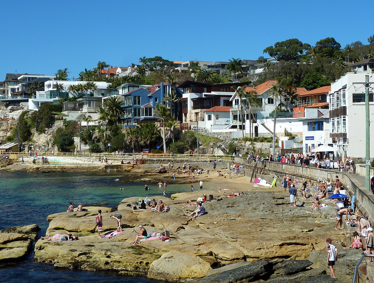 A rock pool cut into the shoreline near Fairy Bower, swimmers and sunbathers on the rocks, houses on the headland behind.