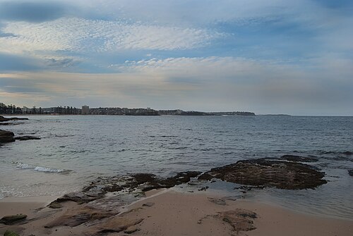 A wide stretch of ocean beach and calm water, the coastline curving away toward distant apartment towers.