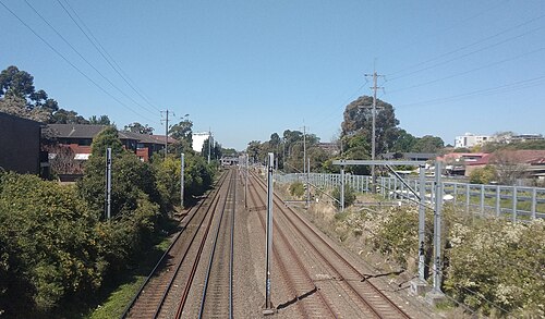 Multiple railway tracks running between overhead wiring gantries, fencing and trees, with rowhouse rooftops alongside.