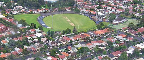 An aerial view of Henson Park, a sunken sports oval with a grandstand along one side, ringed by streets of red tile roof houses.
