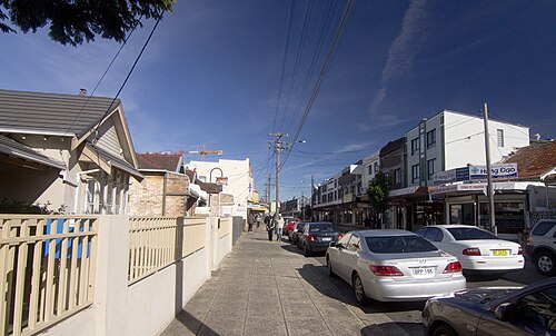 A weatherboard cottage behind a picket fence on Illawarra Road, with Vietnamese shopfront signage visible on the block opposite.