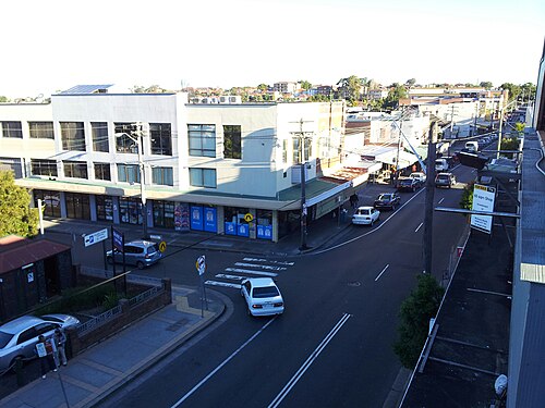 An elevated view of an Illawarra Road intersection, an interwar parapeted commercial building with a For Lease sign, pedestrian crosswalk and parked cars.