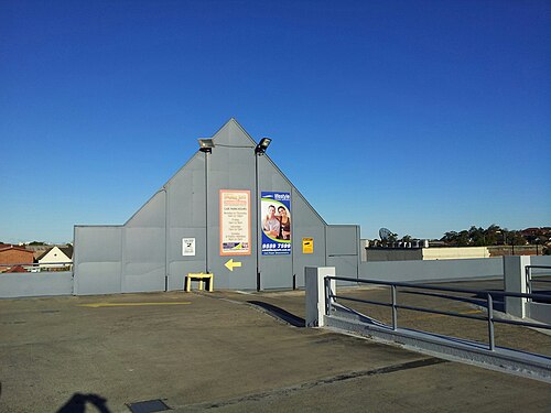 A rooftop car park deck behind Illawarra Road, a grey gabled service wall with floodlights and an advertising panel.