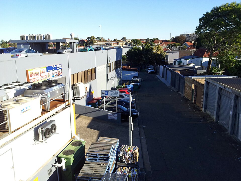 An elevated view down a service laneway behind Illawarra Road, low corrugated roofed sheds, rooftop condenser units and roller doors backing onto the shops. The kind of industrial building Stockade Brew Co occupied, rather than the Cadogan Street building itself, which Commons has no photograph of.