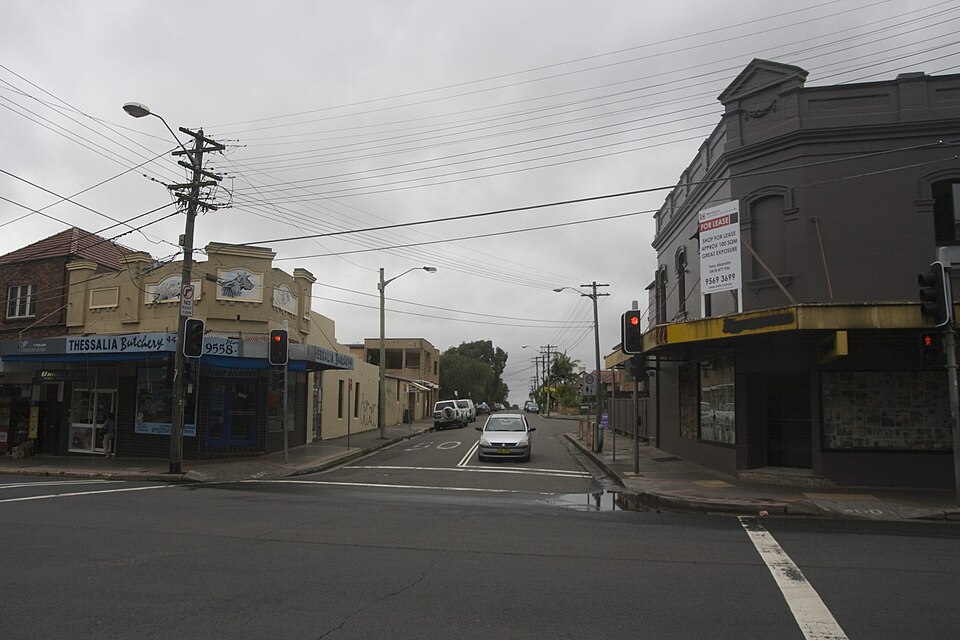 A parapeted corner shopfront on Illawarra Road with an awning over the footpath, traffic signals and terrace shopfronts down the block. The kind of two storey Federation-era corner shopfront the Victoria Road building is, rather than the building itself, which Commons has no photograph of.