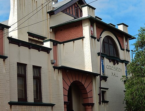 The gabled brick front of Coronation Hall, an arched window over the entrance and CORONATION HALL lettered in relief.