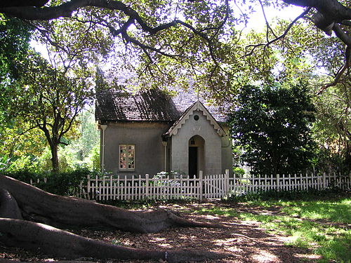 The Camperdown Cemetery lodge, a small Gothic gatehouse in render with a steep gable, behind a white picket fence under fig trees.