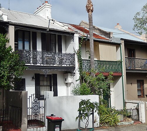 A pair of two storey terrace houses with cast iron lace balconies, corrugated iron roofs and chimney pots behind painted parapets.