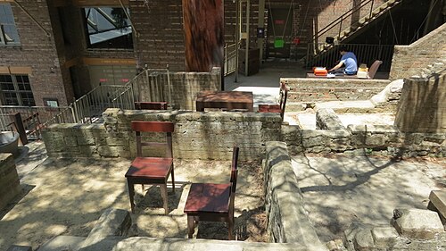 Excavated sandstone footings and low rubble walls stepped into a courtyard behind a brick warehouse, garden chairs set among the ruins.