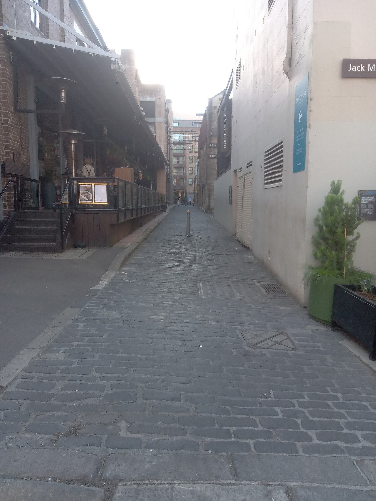 A cobbled lane running between a cafe under an awning and a plain rendered wall, terraces visible at the far end.