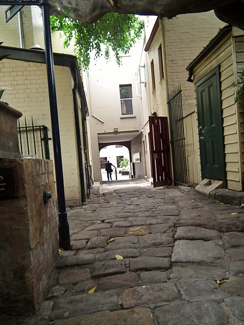 A cobbled lane between a cream terrace and a corrugated iron fence, an open timber gate and a figure walking through an archway at the far end.