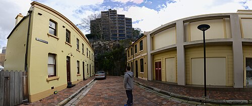 Yellow rendered terraces on a bending cobbled lane below an exposed sandstone rock face, a modern apartment block rising directly above it.