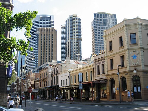 A row of sandstone and rendered shopfronts including a pizzeria and a police station under a bank of high rise towers, a stop sign at the corner.