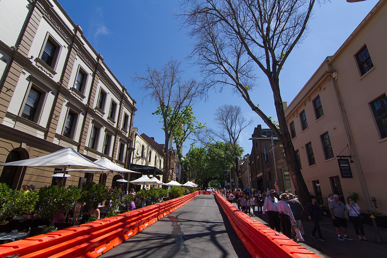 A wide street of two and three storey rendered and masonry terraces on both sides, outdoor tables under umbrellas, plane trees and orange event barriers down the centre of the road.