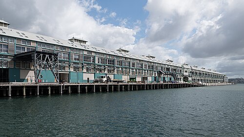 The full length of the Finger Wharf at water level, its timber cladding and corrugated roof running on piles over the harbour.