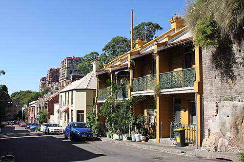 A row of two storey painted terraces with iron lace balconies on McElhone Street, a sandstone rock face behind them and a public housing tower rising further up the hill.