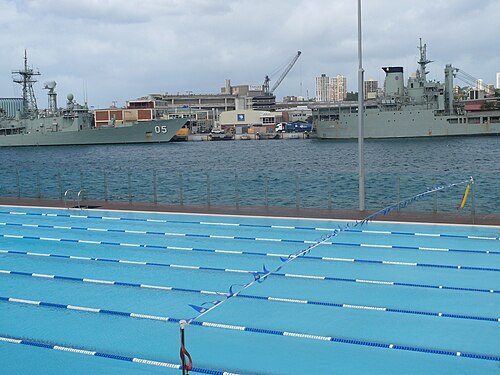 Andrew (Boy) Charlton Pool's outdoor lap lanes on the harbour edge, two grey navy ships moored directly across the water behind the fence.