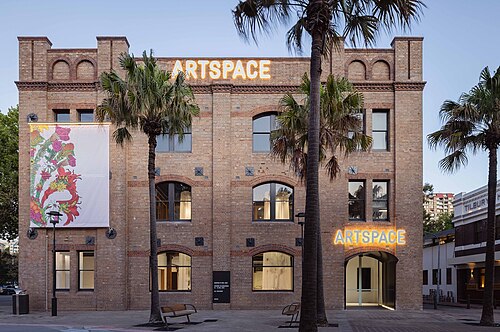The Gunnery's brick facade with arched windows and the lit ARTSPACE sign above the entrance, palm trees along the footpath.