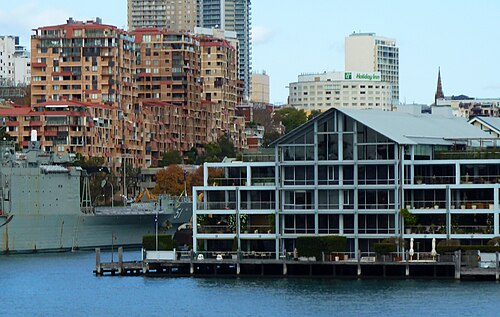 A navy vessel docked at the wharf at close range, seen from across the water with a distant church spire on the skyline behind.