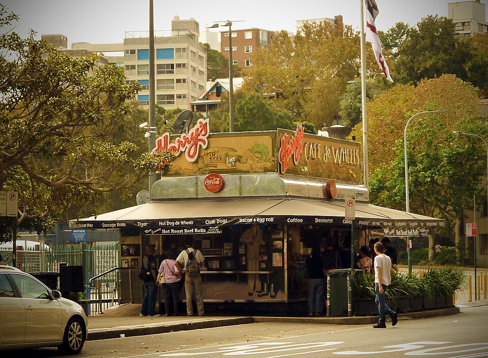 Harry's Cafe de Wheels on its corner, the neon Harry's Cafe de Wheels sign lit above the serving hatch, customers standing at the counter and public housing towers behind.