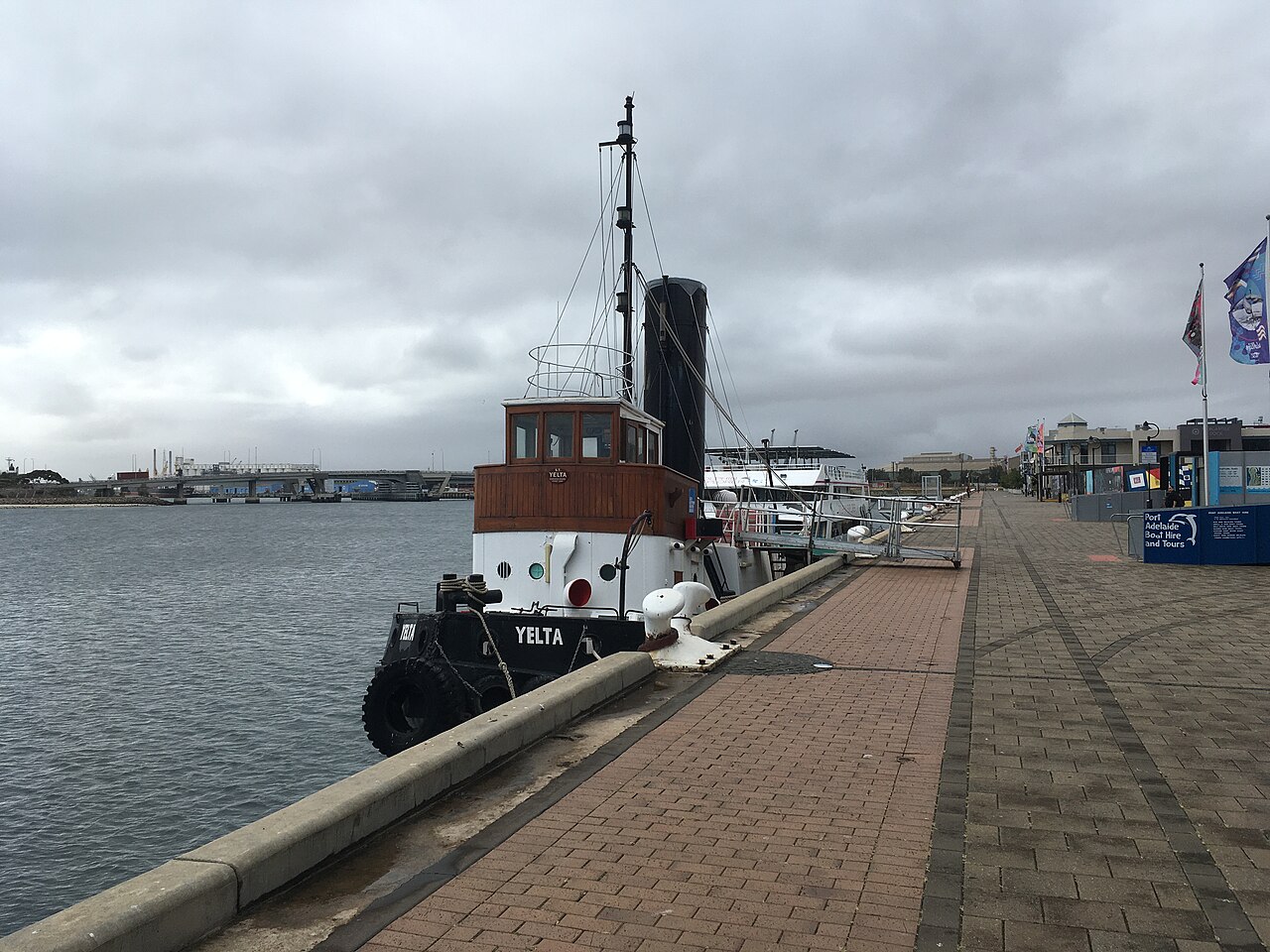 The tug Yelta moored at Queen's Wharf. The brick paved wharf runs off to the right past flagpoles and a boat hire shed, with a bridge in the distance.