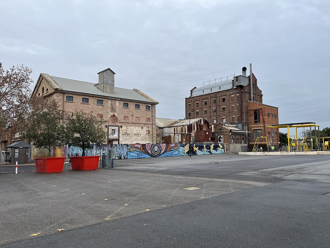Hart's Mill seen across the yard. A stone shed with a corrugated roof stands on the left and the taller brick mill on the right, with rusted iron sheds between them and two skips out on the asphalt.