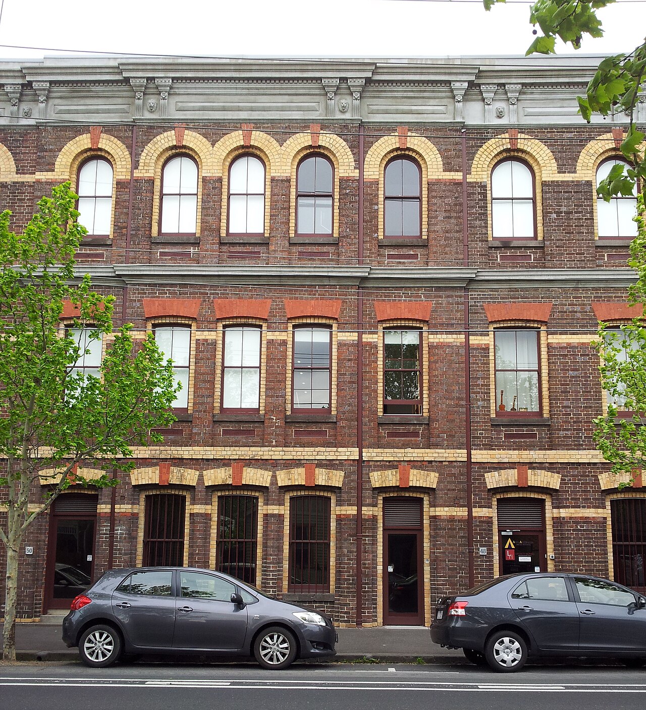 Denton Hat Mills' three storey polychrome brick facade on Nicholson Street, arched upper windows banded in cream brick, cars parked at the kerb.