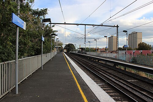 Victoria Park railway station's platform, tracks running under overhead wires with housing towers on the skyline beyond.