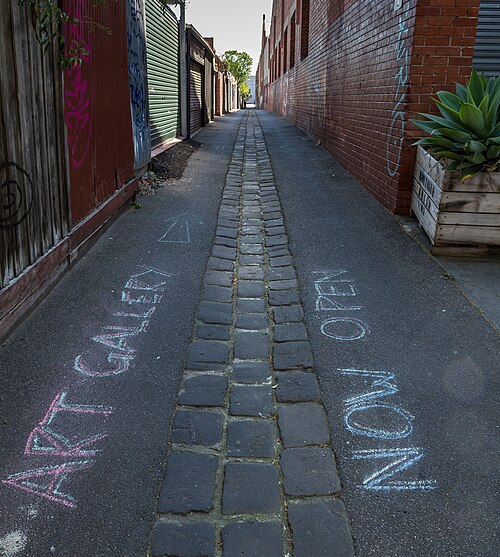 A narrow rear laneway of brick walls and corrugated roller doors, a bluestone cobblestone gutter running down the centre.