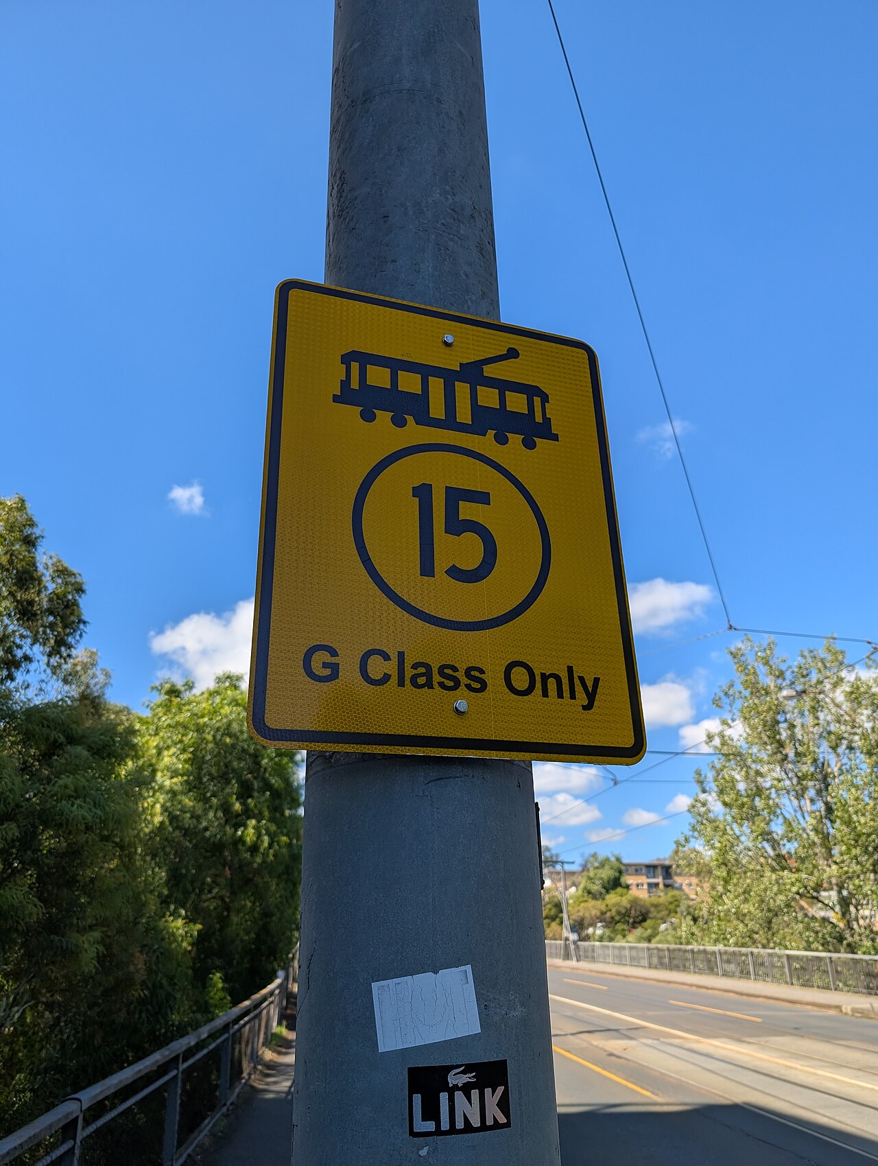 A yellow tram speed limit sign for G class trams on a pole at the west end of Victoria Bridge, the road and railings behind it.