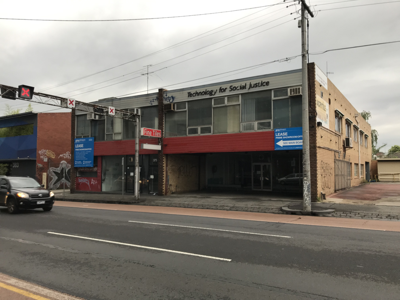 A two storey commercial building on the main road, tan brick and glass shopfronts under a lease sign, tram wires overhead.