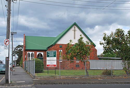 A single storey red brick church with a green corrugated roof, behind a chainlink fence on a side street off Sydney Road.