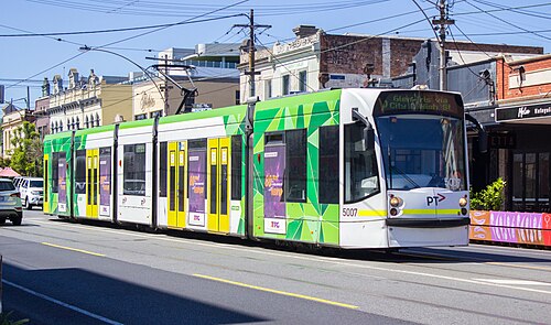 A green and white tram running down Lygon Street, Brunswick, under the overhead wires past a row of shopfronts.