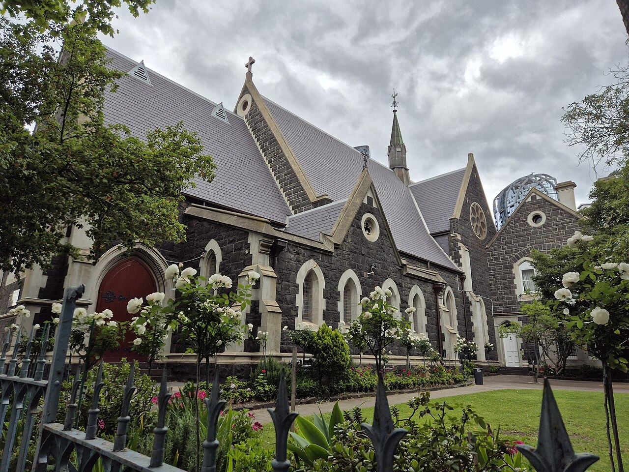 The Romanian Orthodox Church on Queensberry Street, a bluestone Gothic Revival building with a steeply pitched slate roof, arched windows and a garden of white roses behind a wrought iron fence.