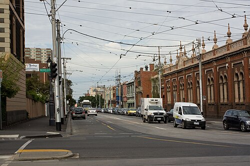 A wide street with overhead tram wires, red brick heritage buildings on both sides and moving traffic under an overcast sky.