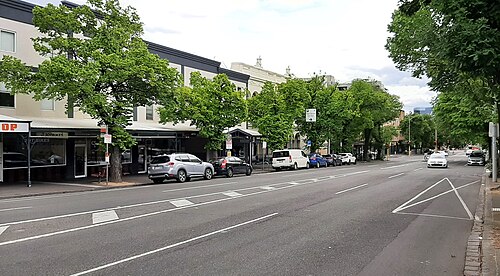Grattan Street, a wide asphalt road with leafy street trees, parked cars and low rise commercial shopfronts under verandahs.