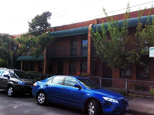 Two storey red brick townhouses with dark green corrugated awnings, cars parked along the kerb.