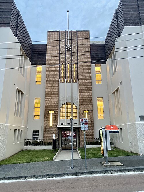 The Collingwood Yards facade on Johnston Street, an Art Deco brick and rendered front with a central arch and a public phone booth on the lawn.