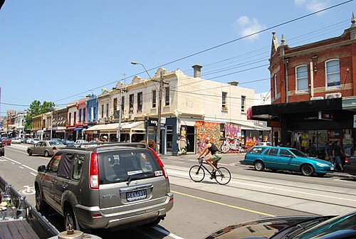 A cyclist riding past two storey Victorian shopfronts in Fitzroy, street art on the walls, parked cars and tram tracks in the road.
