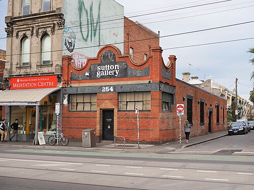 A red brick commercial building with a decorative parapet next to a grey painted shopfront on Brunswick Street, tram tracks in the road.