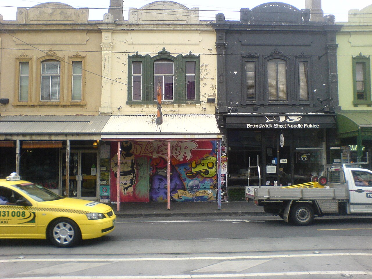A row of two storey Victorian shopfronts in Fitzroy, verandah posts over the footpath, painted facades, street art on an end wall, and a taxi passing.