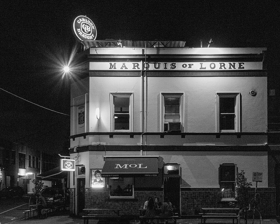 A two storey corner pub lit up at night, painted facade, illuminated signage over the door and outdoor tables at the kerb.