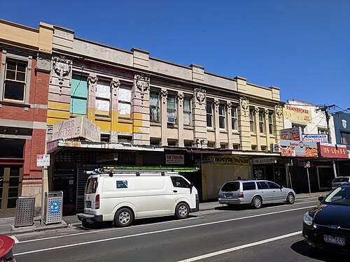 A row of two storey period commercial shopfronts along Barkly Street, parked cars and awnings at the kerb.
