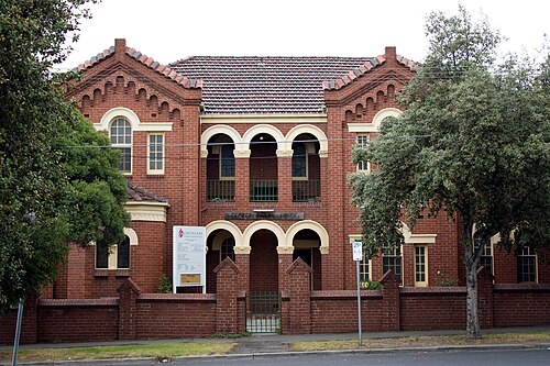 A two storey red brick building with arched loggias and decorative brickwork behind a low brick wall.